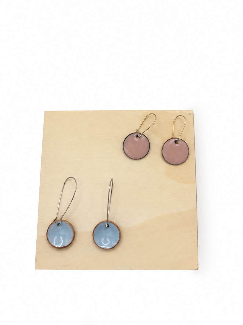 Three pairs of minimalist disc earrings displayed on a light beige square card against a solid white background. The earrings feature circular flat tops in light blue and dusty rose, suspended from thin gold-toned wires.